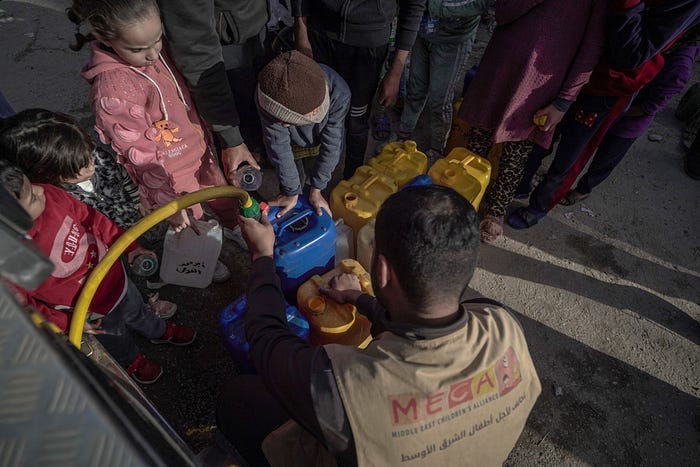 MECA staff filling up jugs of clean drinking water for displaced families in Rafah, Gaza. The photo shows a man wearing a MECA vest filling a jug for one child, with three other children holding jugs, while several adults wait in line in the background. Used with permission from MECA.