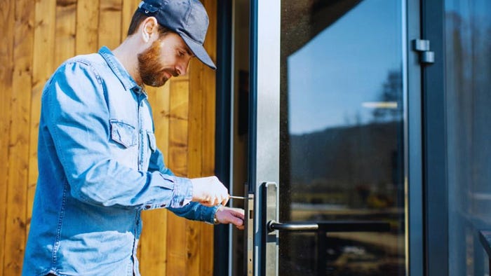 A man in a denim shirt and cap installs a modern glass door, using a tool to adjust the exterior handle on a wood-sided home.