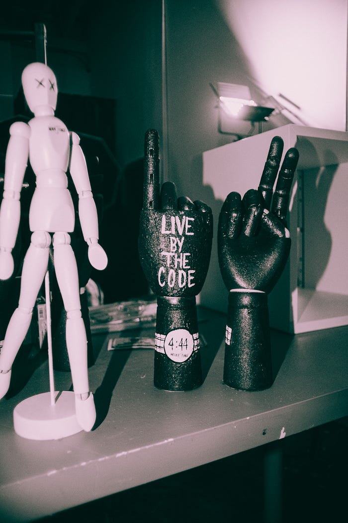 A black and white photograph of a mannequin and two mannequin hands, one of which has a raised index finger with the words “Live by the code” written on it.