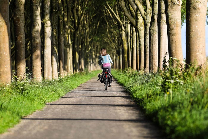 A women going away from the camera with her bike, while peddaling alongside trees.