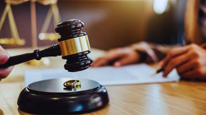 Close-up of a judge's gavel next to a wedding ring on a sound block, with a person signing divorce documents in the background, symbolizing legal proceedings and marital separation.