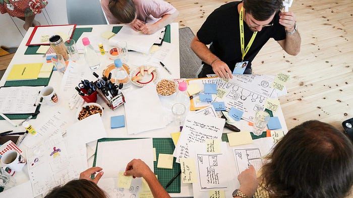 A birds-eye-view of a group of people working on creating storyboards with sticky notes. There are papers and office supplies covering the tables.
