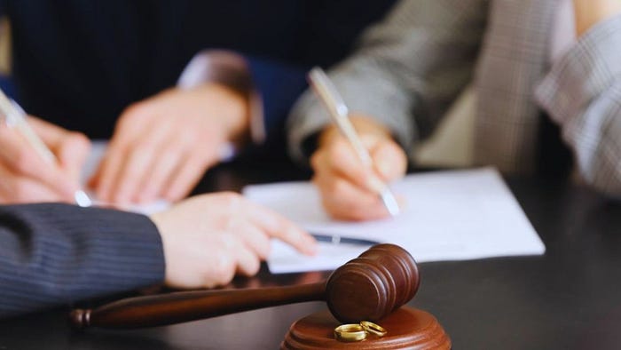 Close-up of two people signing divorce papers at a desk, with a judge’s gavel and a pair of wedding rings in the foreground, representing legal involvement in a divorce case with a lawyer’s guidance.