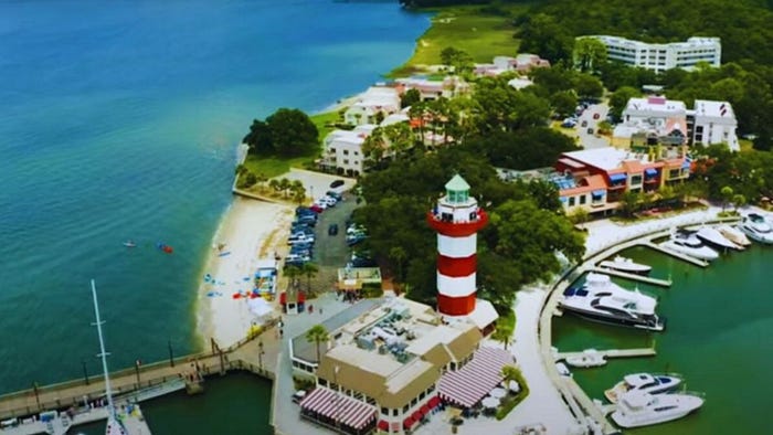 Aerial view of Hilton Head Island, SC harbor featuring boats and a lighthouse surrounded by calm waters.
