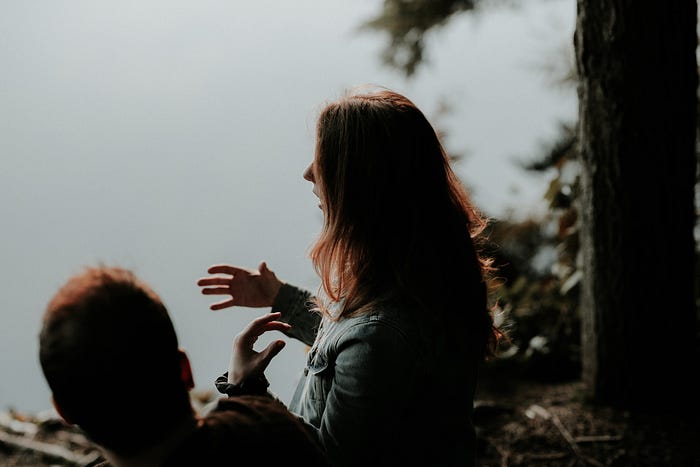 A woman with medium length brown hair stands beside a man overlooking a lake, talking with her hands