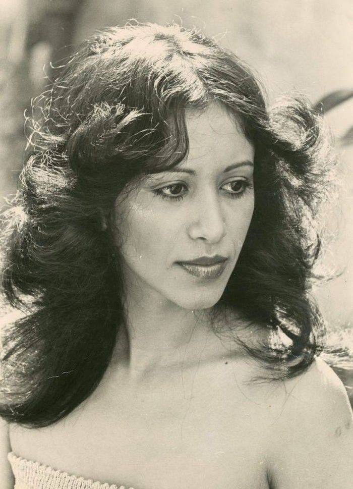 Liat Portal for The Liat Show: A black-and-white portrait of Israeli singer Ofra Haza in her youth. She wears a strapless textured top, her long wavy hair framing her face as she gazes downward with a reflective, almost shy expression. The natural light highlights her delicate features and quiet charisma, capturing the early promise of a voice that would later carry Yemenite tradition and modern pop to the world stage.