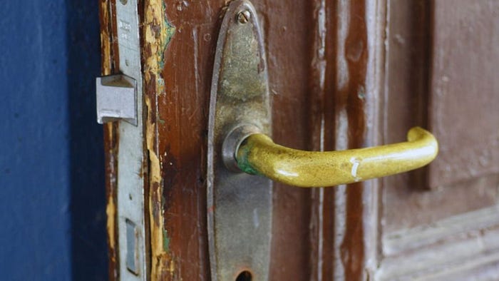 A close-up of a worn wooden door with peeling paint and a tarnished brass handle, showing visible damage and corrosion around the lock and handle area.