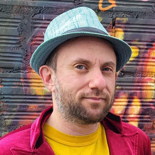 Headshot of Tucker Lieberman in a bright yellow T-shirt and magenta velvet jacket. He is a 40-ish white man with blue eyes and a graying beard. He’s wearing a blue sun hat with a brim and hatband to hide his baldness. Behind him is a graffiti-covered concrete wall.