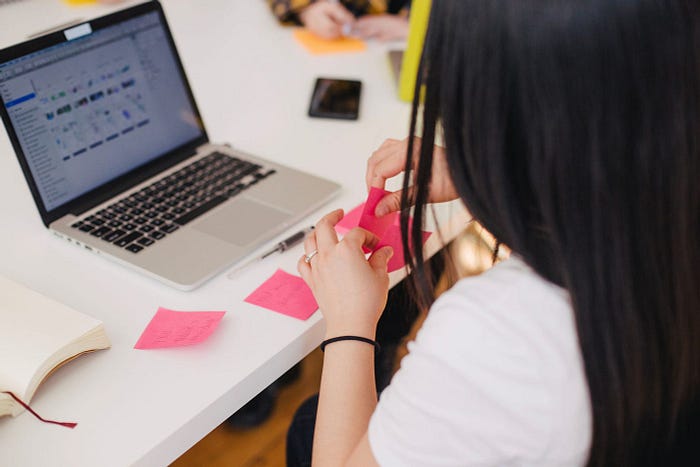 A close up of a person holding a pink sticky note while in front of a laptop. Here are other pink sticky notes on the table in front of her.