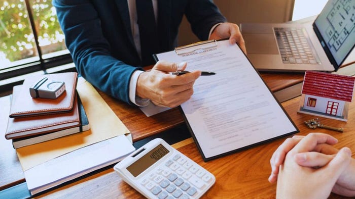 A real estate agent in a business suit reviews paperwork with a client at a wooden desk, which also holds a laptop, calculator, notepads, a house key, and a small model of a house, in a well-lit office environment on Hilton Head Island, SC.