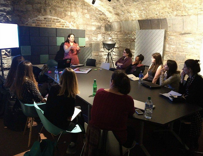 A woman speaks to a group of women sitting at a conference table in a stone-walled room.