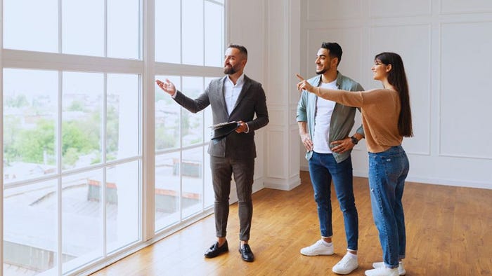 Young couple viewing a bright, empty apartment with a real estate agent standing by large floor-to-ceiling windows, discussing features of the modern hardwood-floored space.