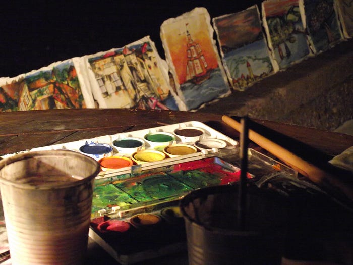 A colourful palate with a paintbrush and water cup rests on a table before painted tiles of a boat, a lighthouse, and scenes of the sea.