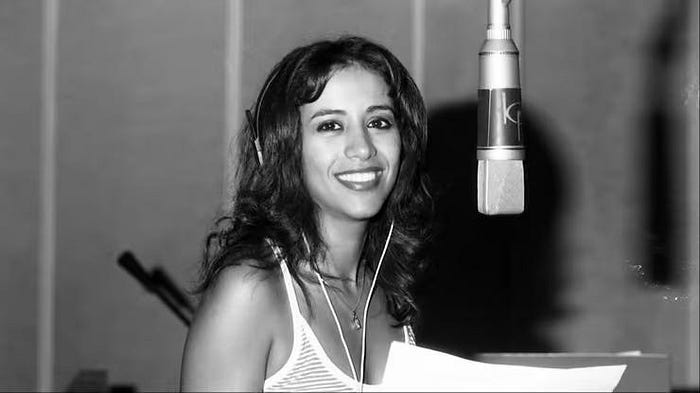 Liat Portal for The Liat Show: A black-and-white photograph of Israeli singer Ofra Haza smiling while recording in a professional studio. She wears headphones and a striped tank top, holding sheet music as she stands near a large condenser microphone. This moment reflects her creative process and international success, where her Yemenite roots fused with pop production to create a timeless global sound.