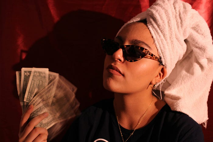 A young woman with sunglasses holding a spread of dollar bills. She’s wearing a pink towel turban.