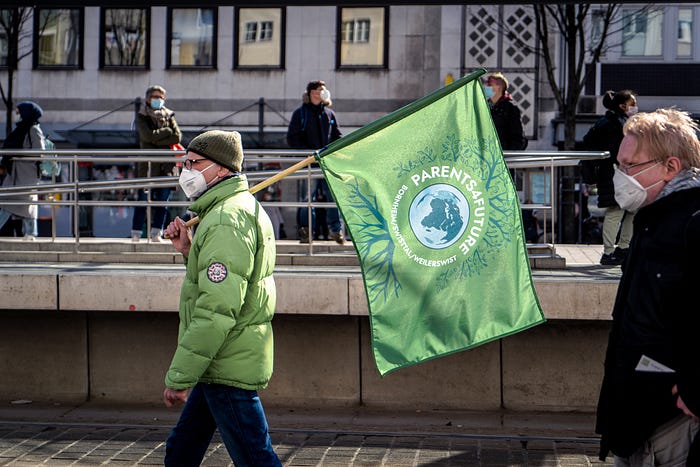 A person in a green jacket holding a green flag that reads:”PARENTS 4 FUTURE”.