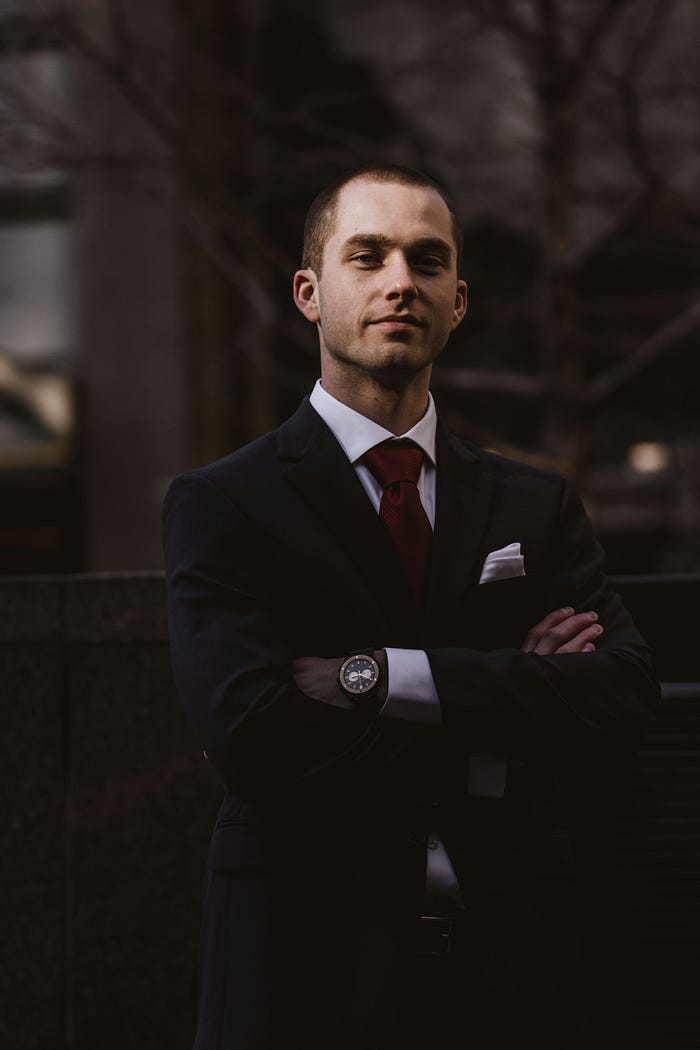 A young man in a suit, standing outdoors, arms folded on his chest, looking fulfilled