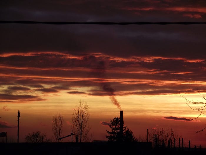 Smoke stacks letting off fumes into the sky, against the backdrop of a bloody sunset and ominous clouds.