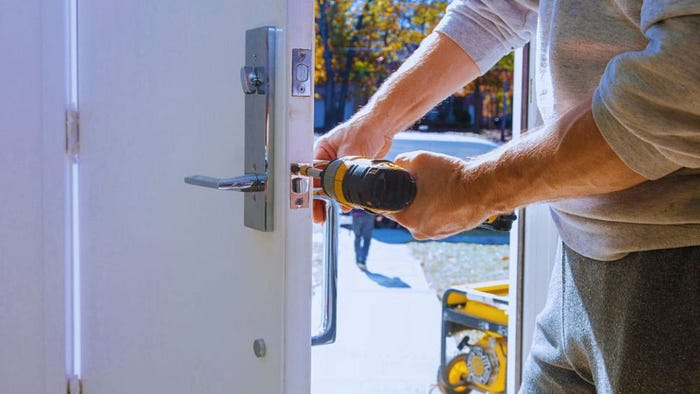 A professional door installer uses a power drill to secure a modern metal lock on a white exterior door during daylight.