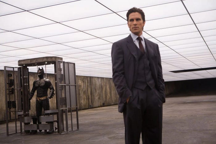Christian Bale sebagai Bruce Wayne di film Batman Begins, berpose di sebelah Batman suit miliknya di sebuah gudang rahasia