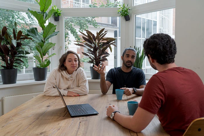 Three people sitting at a table talking. There is a laptop and coffee cups on the table, and plants in the background. They are sitting by a window.