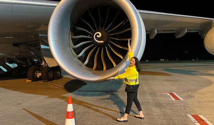 A girl wears a yellow safety vest and poses for a photo with an airplane engine.
