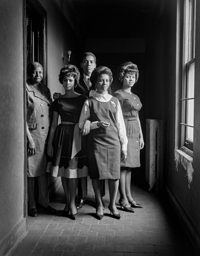 a group emerges from a doorway on the left into a hall with brick floors. Three women are in dress uniform, another is in a fancier dress, and a man in the back wears a suit.