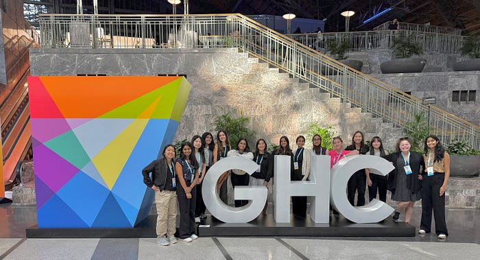 Students stand around large "GHC" letters with a colorful geometric statue and staircase in the background.