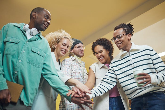 A group of 5 people, different genders and ethnicities placing their hands on top of each other, smiling.