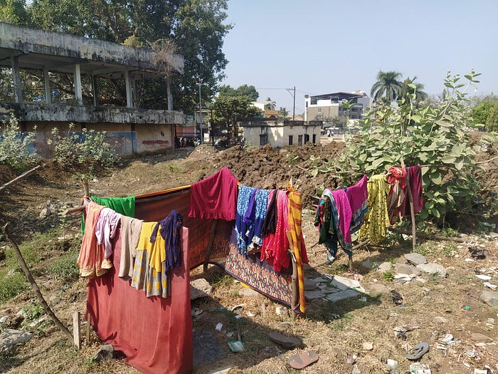 A photo of a makeshift toilet. There is grass and rocks on the ground, and a concrete building in the background. Colourful materials hang around the toilet to give some amount of privacy.