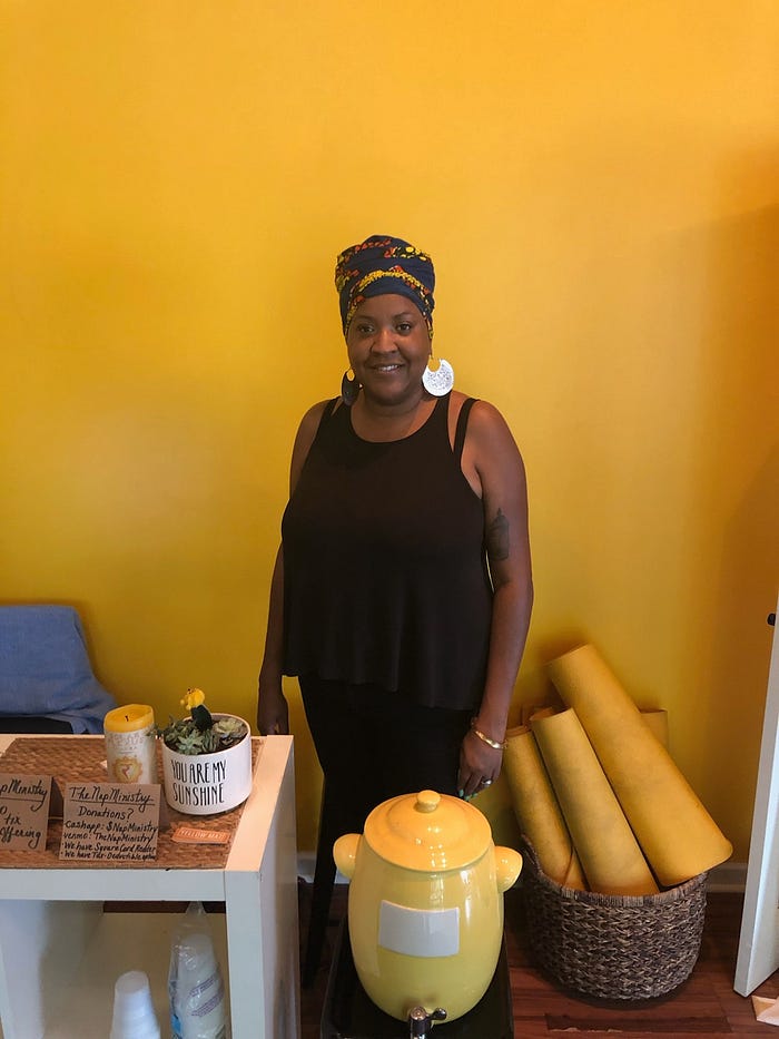 Tricia Hersey, founder of the Nap Ministry, stands against a yellow wall; dressed in black, she stares serenely at the camera.