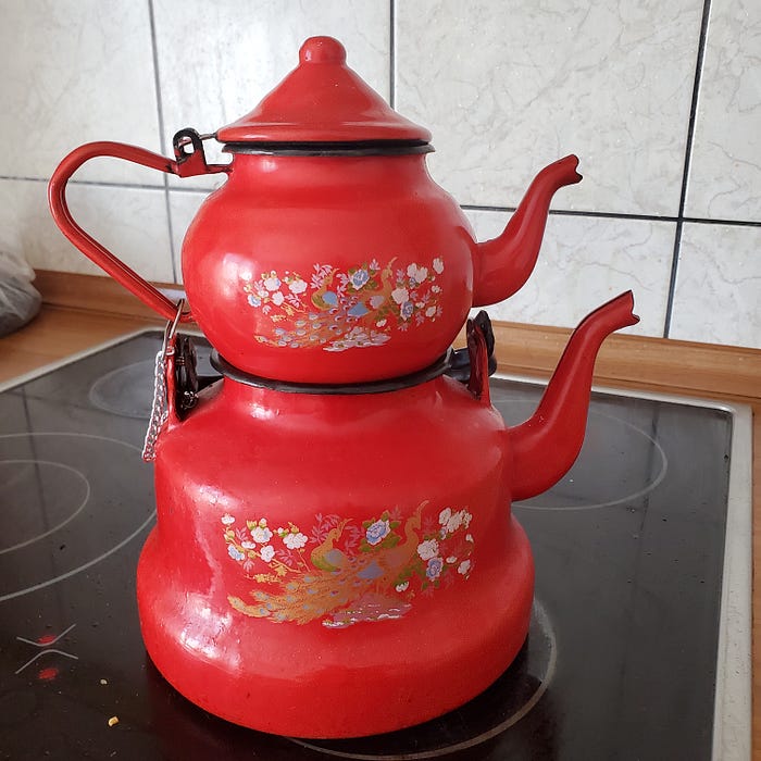 A red enamel double (Turkish style) teapot with a stylized peacock on the front sitting on a modern glass top electric stove.