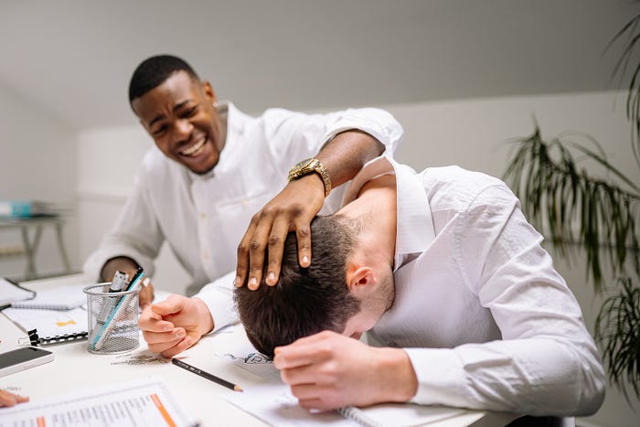 two men wearing white shirts who appear to be office colleagues. One man has his head on the desk, his colleague's hand is on his head. their interaction appears playful.