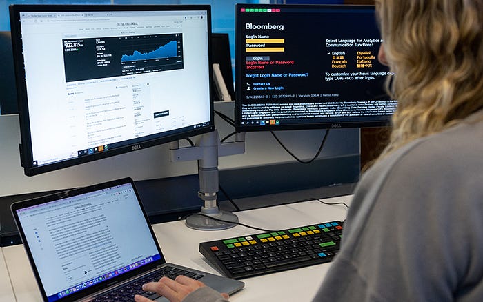 A person is working on a laptop with documentation open, in front of a desktop setup showing Bloomberg financial software on dual monitors.