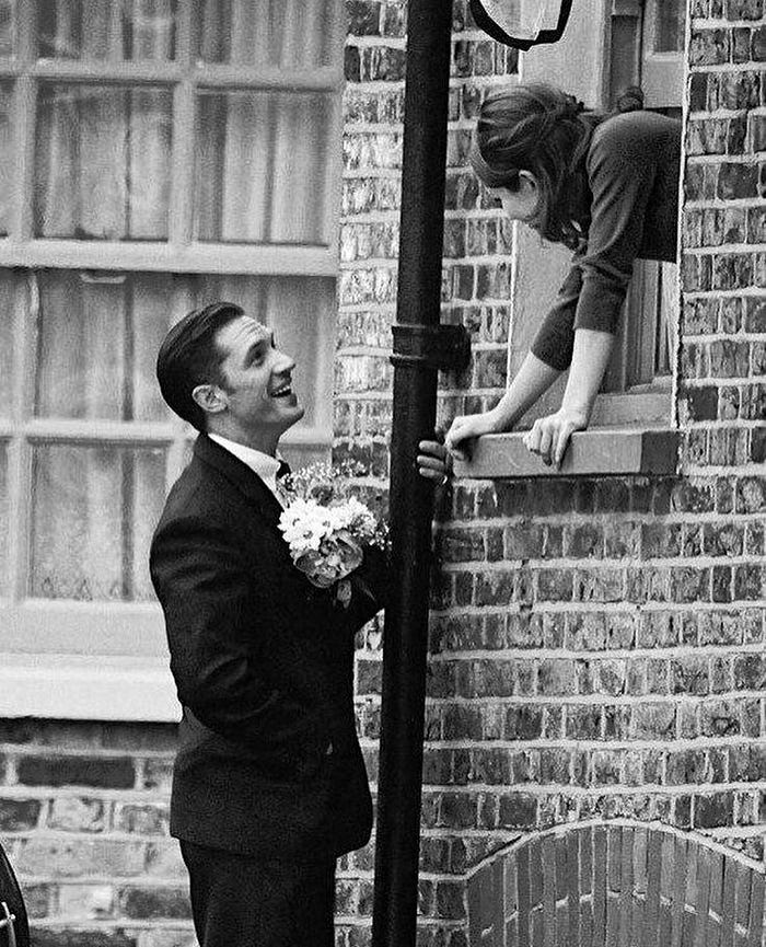 Vintage black-and-white photograph of a man offering flowers to his partner through a window, representing old-fashioned romance before commercial Valentine’s campaigns.