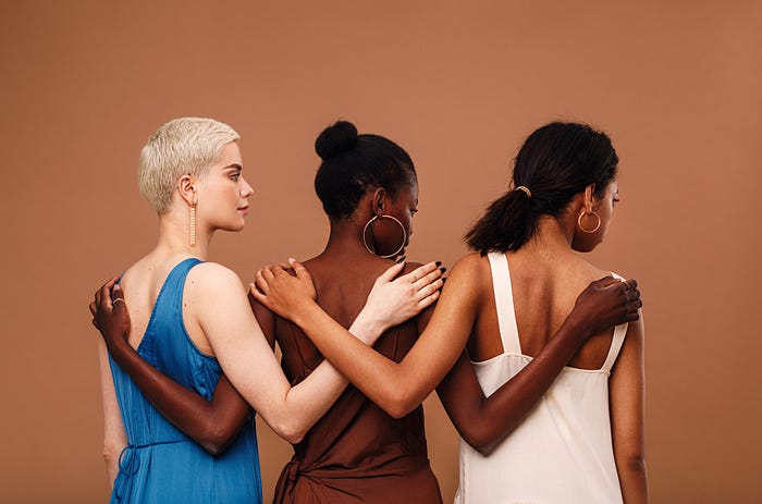 Multi-ethnic women stand with arms on each other’s backs, facing away from the camera, against a light chestnut background.