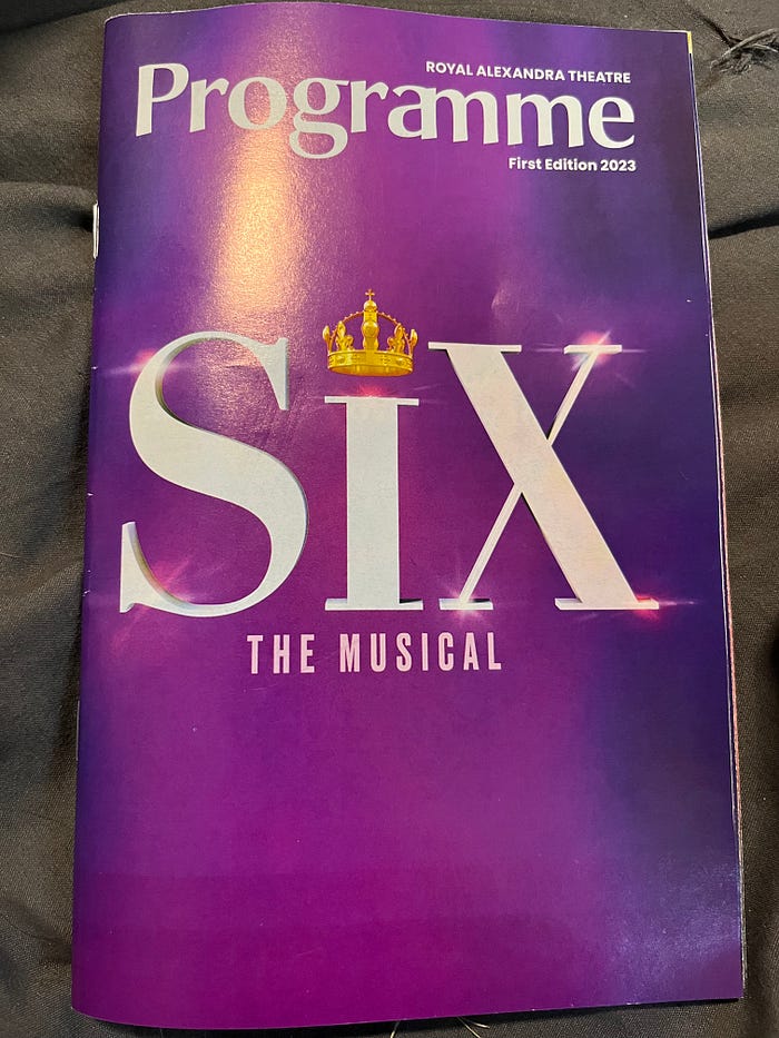 A programme for SIX The Musical. The show???s title is printed in white letters on a purple background, with a gold crown above the I. Additional text in white states that it is a programme, that the show is presented at the Royal Alexandra Theatre, and that it is First Edition 2023.