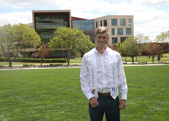 A man wearing a stole reading "student athlete" stands on a lawn overlooking a modern building.