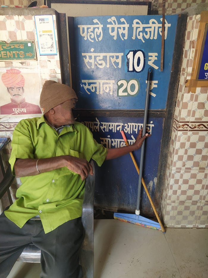 A photo of a man in a green top, with a blurred faced, in front of a blue metal door, and a brown and whit checkered wall. The door has writing on it and with costs for using the toilet, which is behind the blue metal door. The costs are ten and twenty Indian rupees. There is a poster with a cartoon of a man’s face next to the man in the green top.