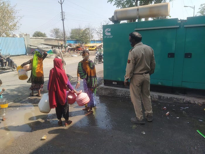 A photo with four people standing outside. Three are women wearing colourful clothing. They are walking away from the camera holding buckets in their hands. The fourth person is a security guard who is blocking them. They stand next to a grey road and a large green generator.