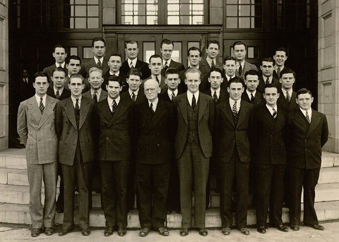 A black and white photo of men dressed in suits standing on stairs leading up to a building.