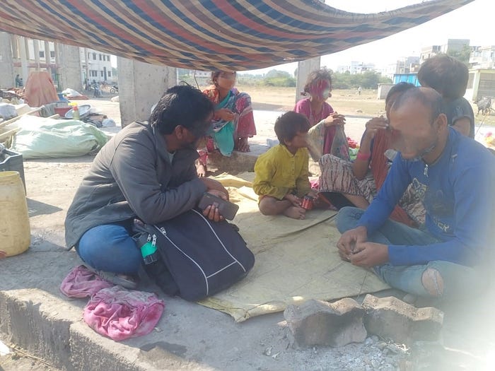 A photo of seven people, including three children, sitting on a pavement. There is a blanket above their heads. Their faces are blurred to protect their identities.