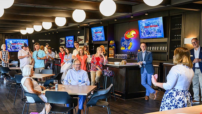 A crowd gathers around a bar with a KU Jayhawk neon sign while facing a female speaker at the front of the room.