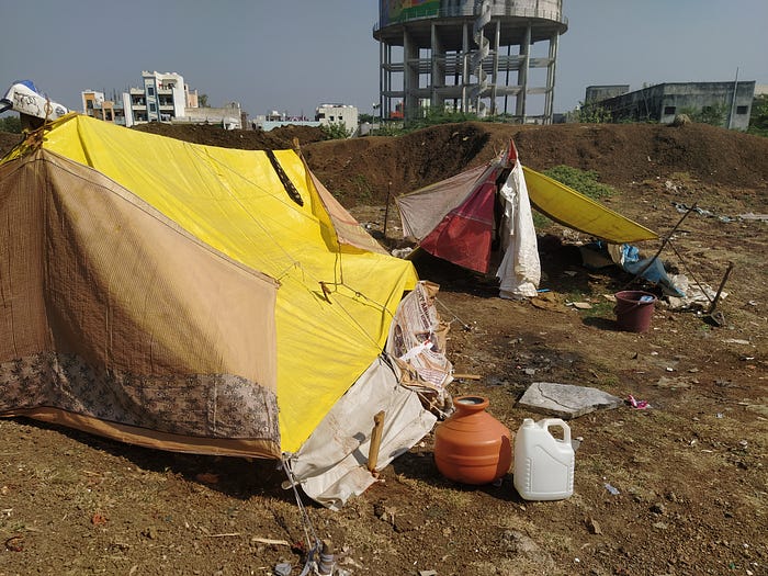 A photo of two makeshift tents, put together with yellow and brown plastic tarps. They stand on top of brown ground, with large concrete buildings in the background. There are some buckets on the ground next to the tents.