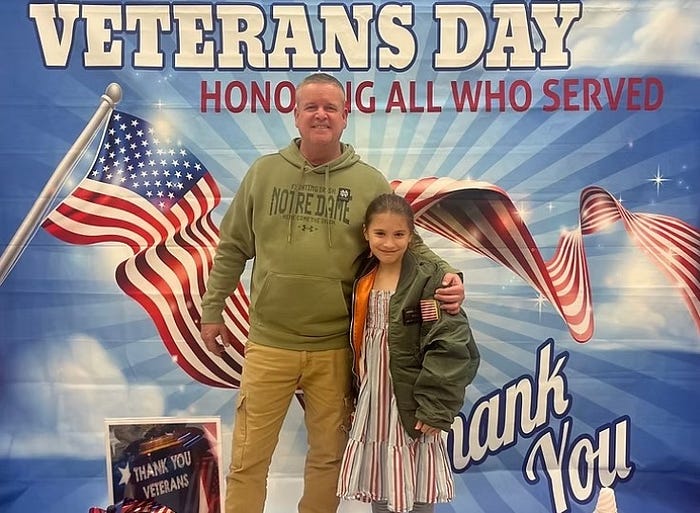 Veteran and child standing in front of a Thank you Veterans Day backdrop.
