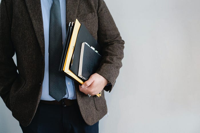 A person in a suit, tie and jacket, holding a bunch of notebooks, but their face is cut off.