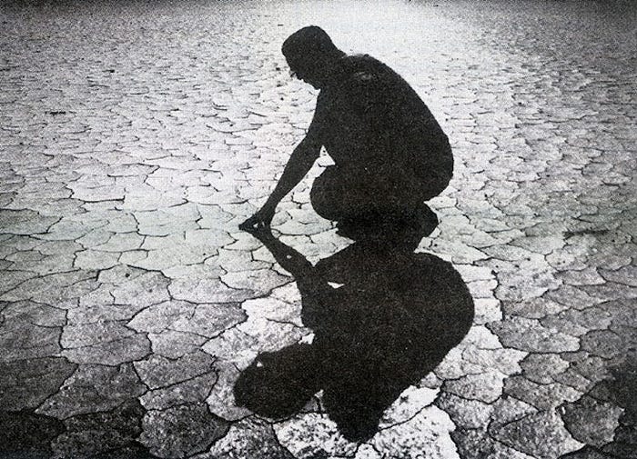 A black and white photo, a man kneels down to touch the ground and the earth is cracked and dry. The sun is in front of the man rendering him in silhouette. His shadow is long and stretches out toward the front of the photo.