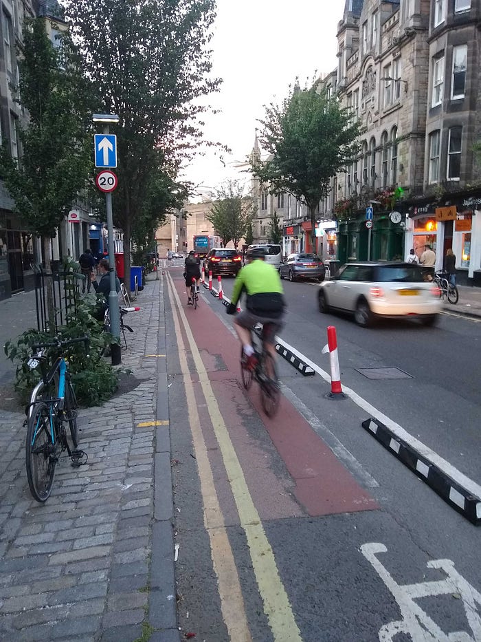 Image shows Forrest Rd in Edinburgh, a narrow one-way street near to the city centre. There are shops & cafes on both sides and the left hand part of the carriageway is coloured red and protected by plastic bollard (or wands) to create a segregated cycle lane. There are two cyclists riding in the bike lane, and several cars driving in the main carriageway. The street is busy with parked vehicles, buses, cars, pedestrians and cyclists.