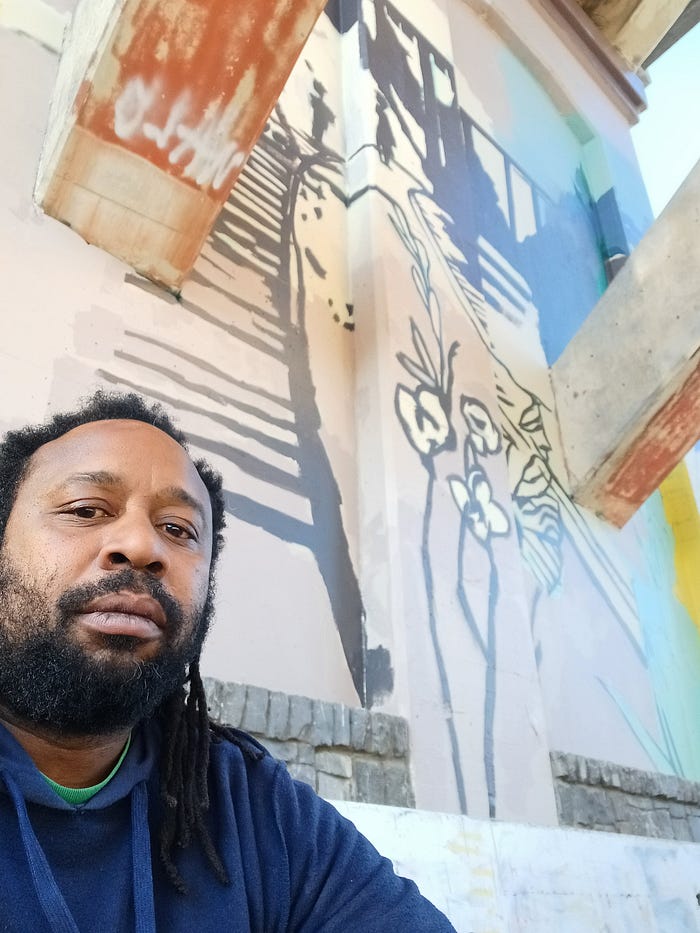 Derrick **Rtystk**Shavers sits in front of a light-colored mural; Derrick has a beard, shoulder-length locks, and a bemused expression.
