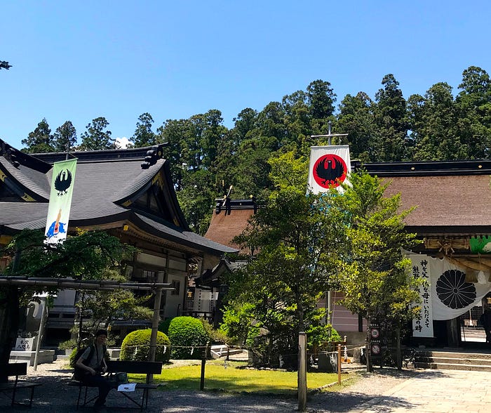 Shrines and a thatched gate, round bushes, trees, and blue sky above. Two banners showing circular, stylized images of a crow with 3 legs.
